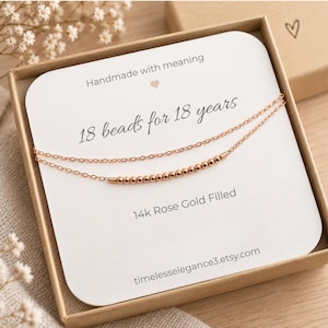 May include: A double-strand rose gold necklace with 18 small beads on a white display card inside a brown box. The card features the text: Handmade with meaning, 18 beads for 18 years, 14k Rose Gold Filled, and timelesselegance3.etsy.com.