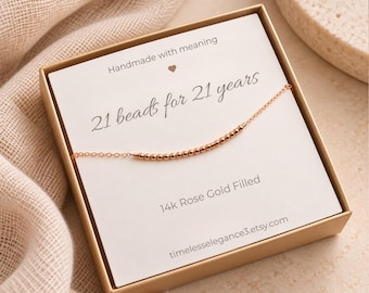 May include: A 14k rose gold filled necklace with 21 small beads on a thin chain, presented in a gold box. The card inside reads: Handmade with meaning, 21 beads for 21 years, 14k Rose Gold Filled, and timelesselegance3.etsy.com.