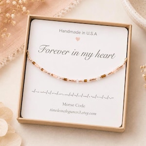May include: A delicate beaded bracelet featuring a Morse code pattern in light pink and metallic brown beads, displayed on a white card inside a cardboard box. The card reads "Forever in my heart" with a small heart icon and Morse code symbols.