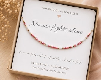 May include: A beaded bracelet with pink, white, and gold-colored beads arranged in a Morse code pattern, displayed on a white card inside a box. The card features the text "No one fights alone" and "Morse Code - 14k Gold Filled".