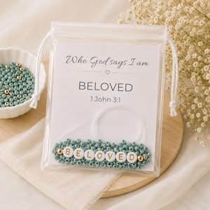 May include: A clear plastic drawstring pouch containing a white card with the text Who God says I am, BELOVED, and 1 John 3:1. Inside the pouch is a coiled clear elastic cord and a row of small sage green beads with white letter beads spelling BELOVED.