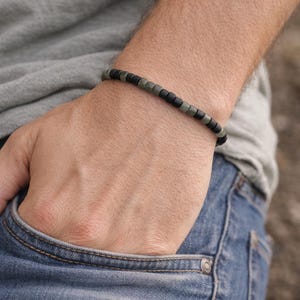May include: A beaded bracelet with alternating black and olive-green square beads. The bracelet is worn on a wrist, with the person's hand in a blue jeans pocket. The person is wearing a gray t-shirt.