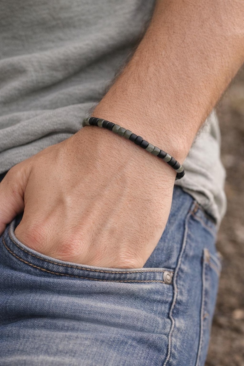 May include: A beaded bracelet with alternating black and olive-green square beads. The bracelet is worn on a wrist, with the person's hand in a blue jeans pocket. The person is wearing a gray t-shirt.