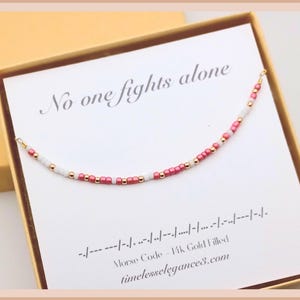 May include: A pink and white beaded bracelet with gold accents. The bracelet is in a box with a white card that reads "No one fights alone" and the Morse code for the phrase. The card also includes the text "Morse Code - 14k Gold Filled" and "timelesselegance3.com".