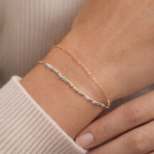 May include: Two delicate gold bracelets. The lower bracelet features small, rectangular, light blue beads interspersed with gold beads. The upper bracelet is a simple gold chain. The bracelets are worn on a wrist.