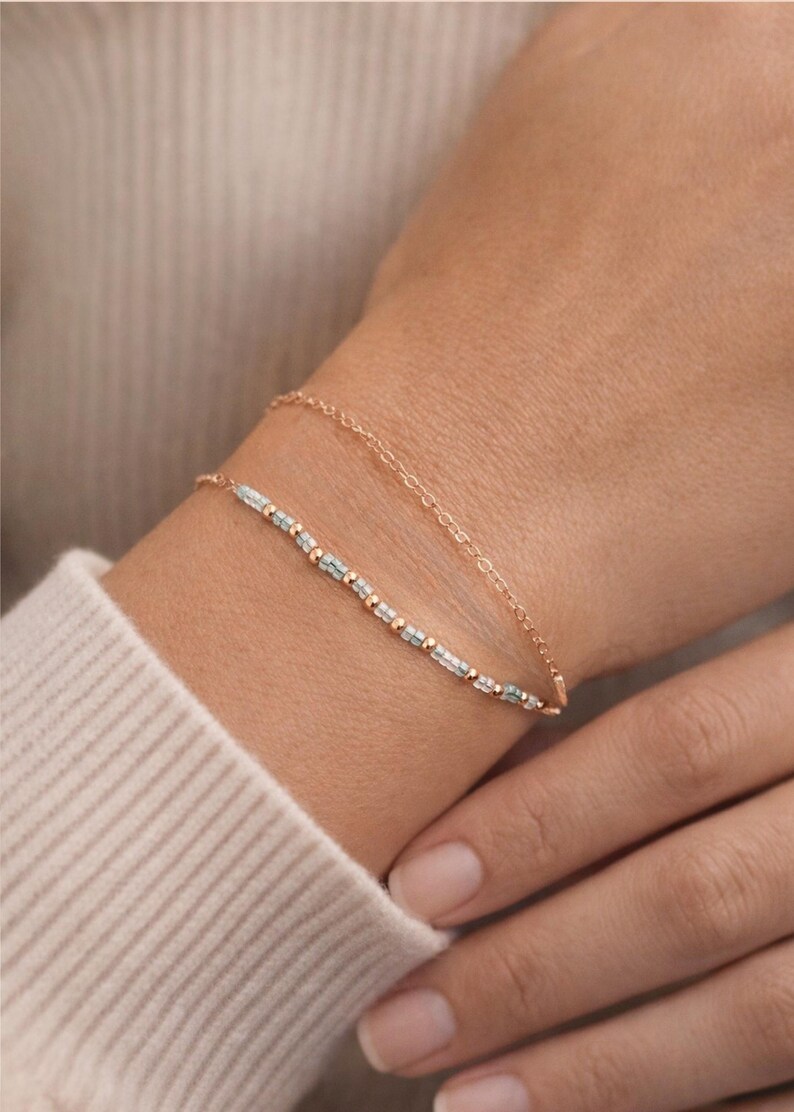 May include: Two delicate gold bracelets. The lower bracelet features small, rectangular, light blue beads interspersed with gold beads. The upper bracelet is a simple gold chain. The bracelets are worn on a wrist.