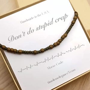 May include: A beaded necklace with a Morse code message, presented on a card that reads "Don't do stupid crap." The necklace features brown and green beads. The card also includes the Morse code translation and the website timelesselegance3.com.
