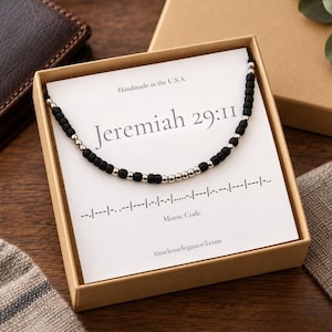 May include: A beaded necklace featuring matte black and metallic silver beads arranged in a Morse code pattern, presented on a white card inside a tan box. The card displays the text Jeremiah 29:11, Morse Code, and Handmade in the U.S.A.