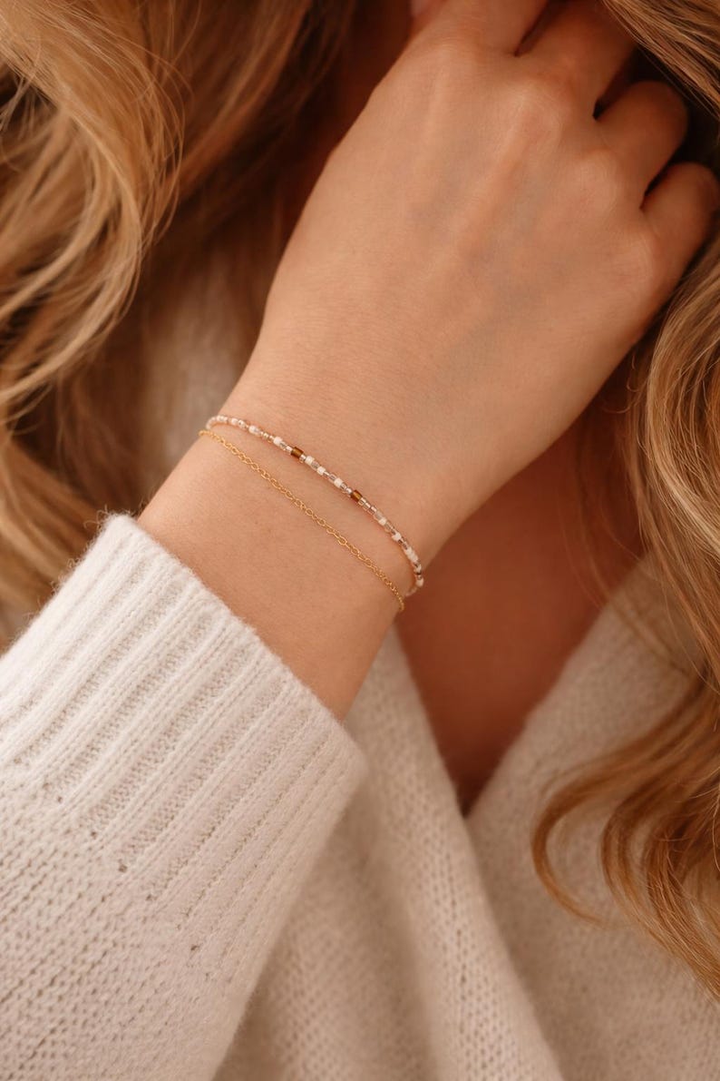 May include: Two delicate bracelets are displayed. The top bracelet features small, light-colored beads interspersed with brown beads. The bottom bracelet is a simple gold chain. The bracelets are worn on a wrist, against a white sweater.