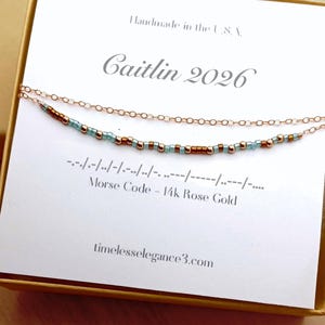 May include: Rose gold necklace with a Morse code design. The necklace features a delicate chain with small beads in teal and brown. The card reads "Caitlin 2026" and "Morse Code - 14k Rose Gold". The website address is timelesselegance3.com.