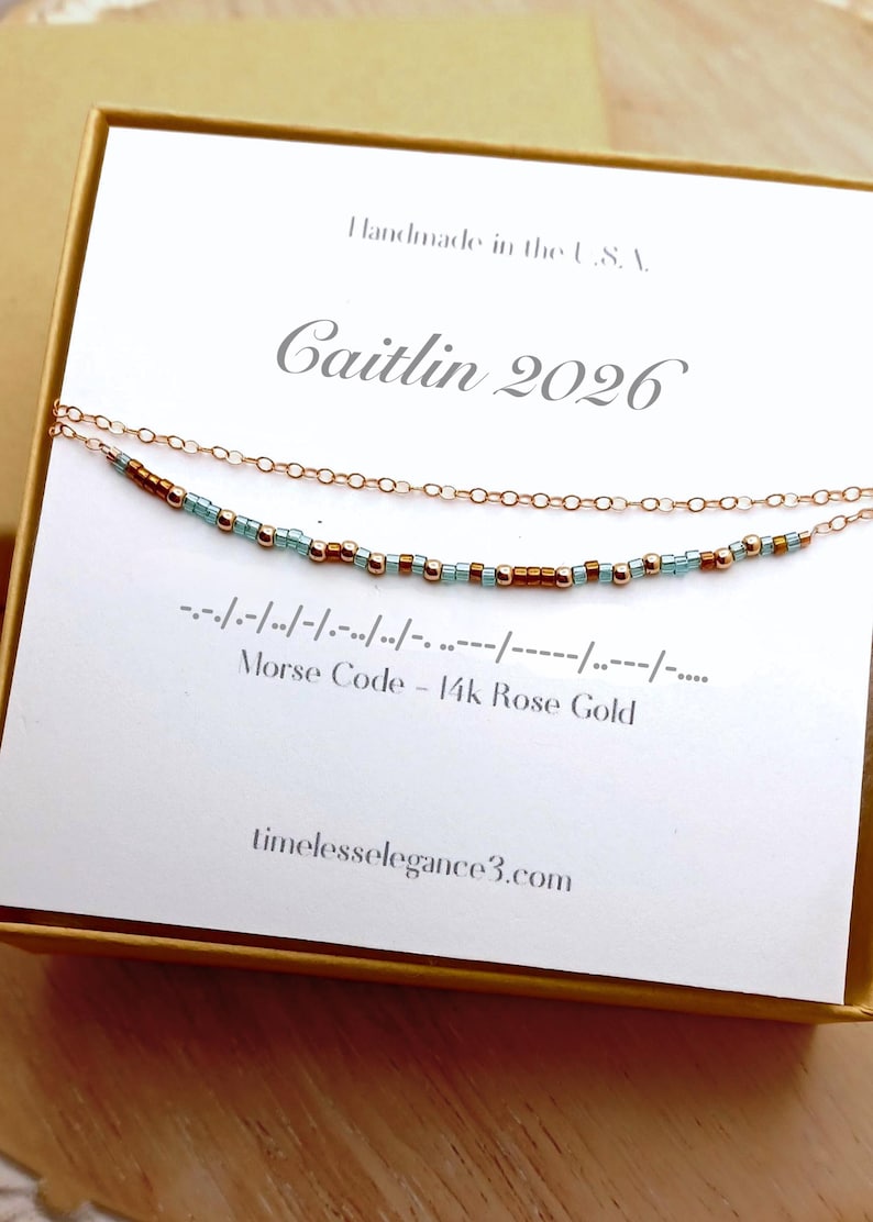 May include: Rose gold necklace with a Morse code design. The necklace features a delicate chain with small beads in teal and brown. The card reads "Caitlin 2026" and "Morse Code - 14k Rose Gold". The website address is timelesselegance3.com.