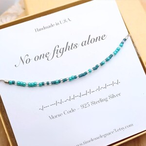May include: A beaded necklace with teal, blue, and silver beads. The necklace is on a white card with the text "No one fights alone" and Morse code. The card reads "Handmade in U.S.A." and "925 Sterling Silver".