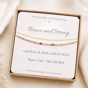 May include: A gold-filled chain necklace with a beaded strand featuring Morse code, presented on a white card inside a brown box. The card displays the text "Handmade with meaning", "Brave and Strong", Morse code symbols, and "Morse Code - 14k Gold Filled".