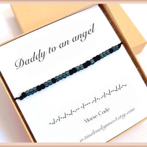 May include: A black cord bracelet featuring alternating matte black and translucent dark green beads arranged in a Morse code pattern. The bracelet is displayed on a white card printed with the text Daddy to an angel and Morse code symbols.