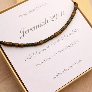 May include: A necklace with a Morse code design, featuring brown and green beads. The necklace is displayed on a card with the text "Jeremiah 29:11" and "Morse Code - 14k Rose Gold Filled". The card is inside a gold box.