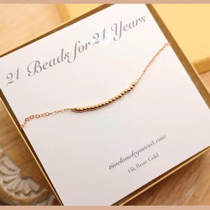 May include: A rose gold necklace with 21 small beads, presented in a gift box. The card inside the box reads "21 Beads for 21 Years" and "timeless elegance3.com". The necklace is delicate and elegant.
