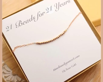 May include: A rose gold chain bracelet with twenty-one small beads, displayed on a white card featuring the text 21 Beads for 21 Years, timelesslegance3.com, and 14k Rose Gold. The bracelet is positioned diagonally across the card.
