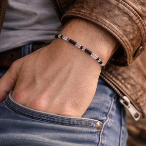 May include: A beaded bracelet with alternating black and clear beads. The bracelet is worn on a wrist, with a brown leather jacket and blue jeans in the background. The bracelet is a simple, minimalist design.