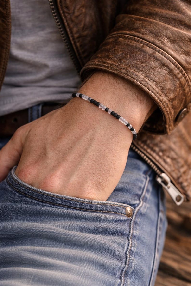 May include: A beaded bracelet with alternating black and clear beads. The bracelet is worn on a wrist, with a brown leather jacket and blue jeans in the background. The bracelet is a simple, minimalist design.