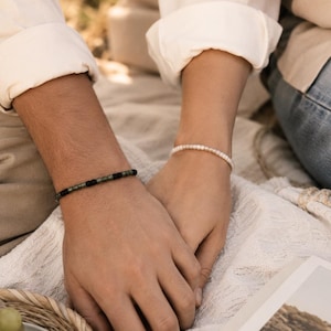 May include: Two beaded bracelets worn on wrists. One bracelet features a repeating pattern of small, cylindrical dark green and matte black beads. The second bracelet consists of a single strand of small, round, iridescent white pearls.