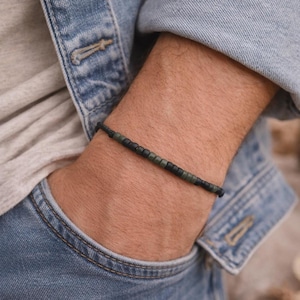 May include: A beaded bracelet with black and olive green beads. The bracelet is strung on a black cord and is worn on a wrist. The person is wearing a denim jacket and a gray shirt.