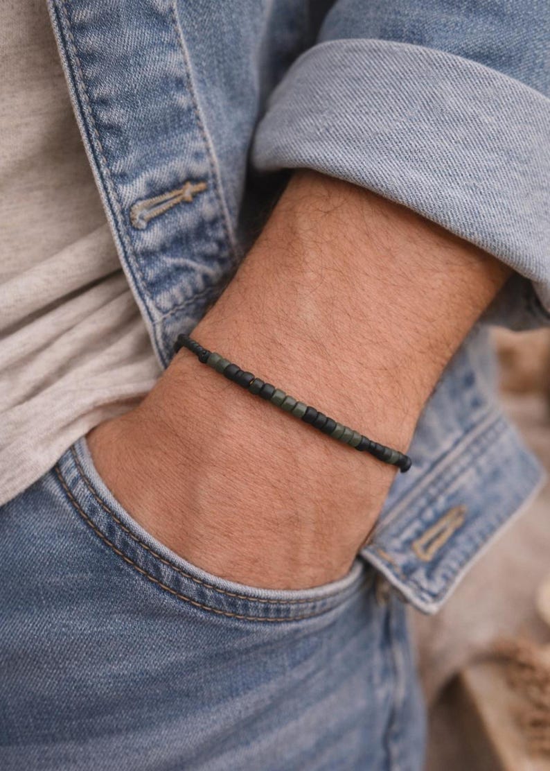 May include: A beaded bracelet with black and olive green beads. The bracelet is worn on a wrist, with a denim jacket and gray shirt in the background. The bracelet is a simple, minimalist design.