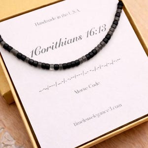 May include: A black and gray beaded necklace presented in a yellow box. The necklace is displayed on a card with the text "1 Corinthians 16:13" and "Morse Code". The box is on a wooden surface.