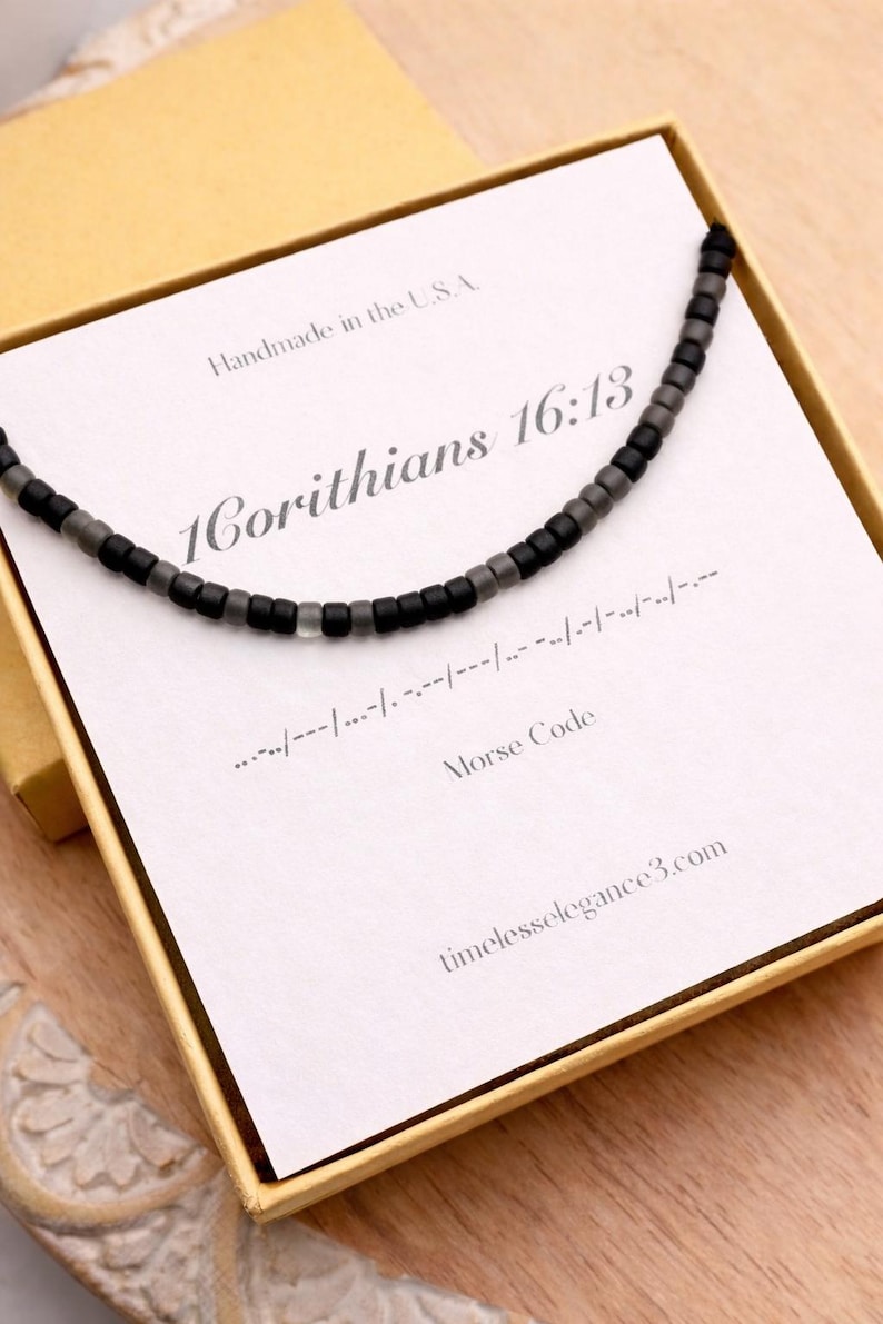 May include: A black and gray beaded necklace presented in a yellow box. The necklace is displayed on a card with the text "1 Corinthians 16:13" and "Morse Code". The box is on a wooden surface.
