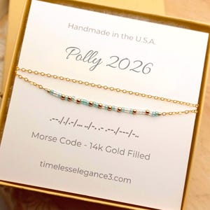 May include: A gold-filled Morse code necklace with light blue and gold beads, presented in a gift box. The card reads "Polly 2026" and "Handmade in the U.S.A." The necklace is delicate and elegant, perfect for gifting.