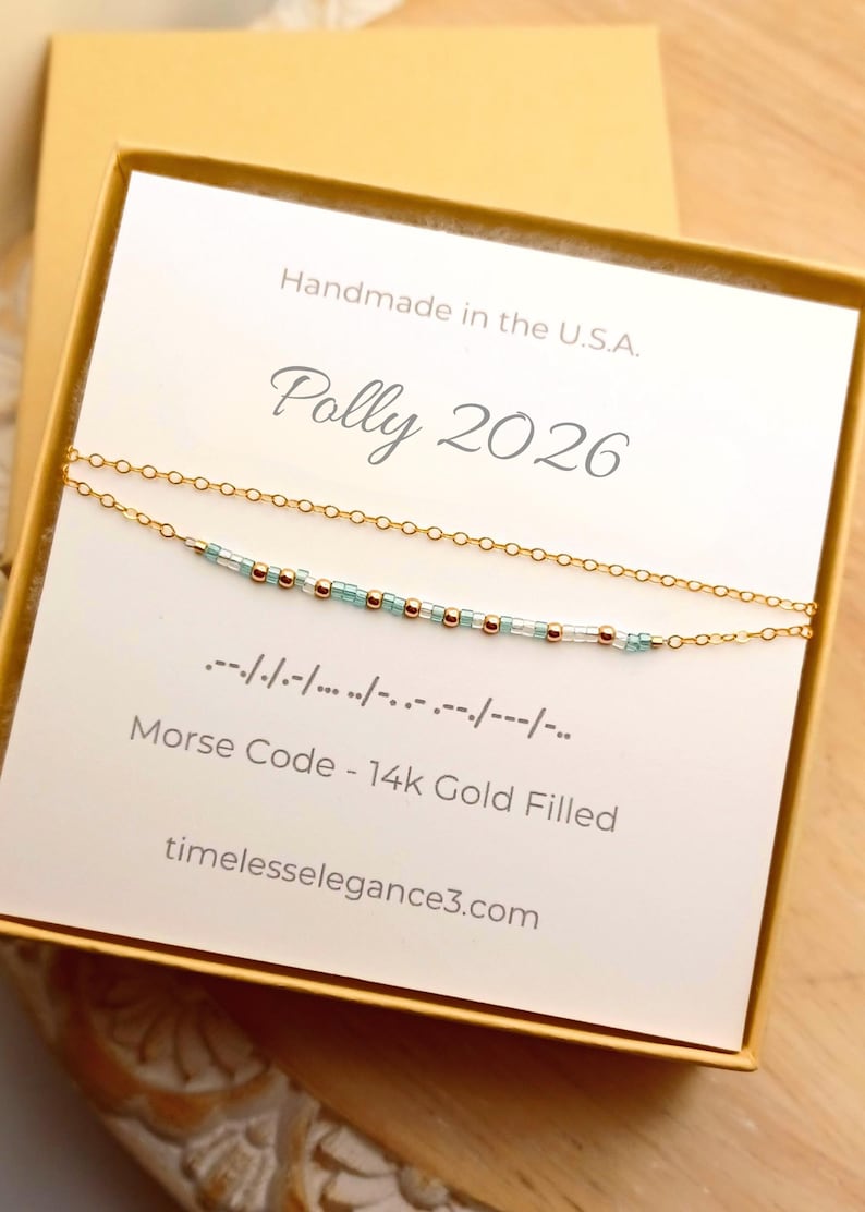 May include: A gold-filled Morse code necklace with light blue and gold beads, presented in a gift box. The card reads "Polly 2026" and "Handmade in the U.S.A." The necklace is delicate and elegant, perfect for gifting.