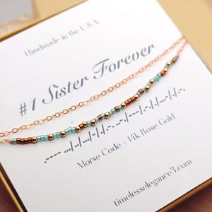 May include: A rose gold necklace with a delicate chain and a beaded section spelling out a message in Morse code. The card reads "#1 Sister Forever" and "Morse Code - 14k Rose Gold". The words "Handmade in the USA" are also visible.