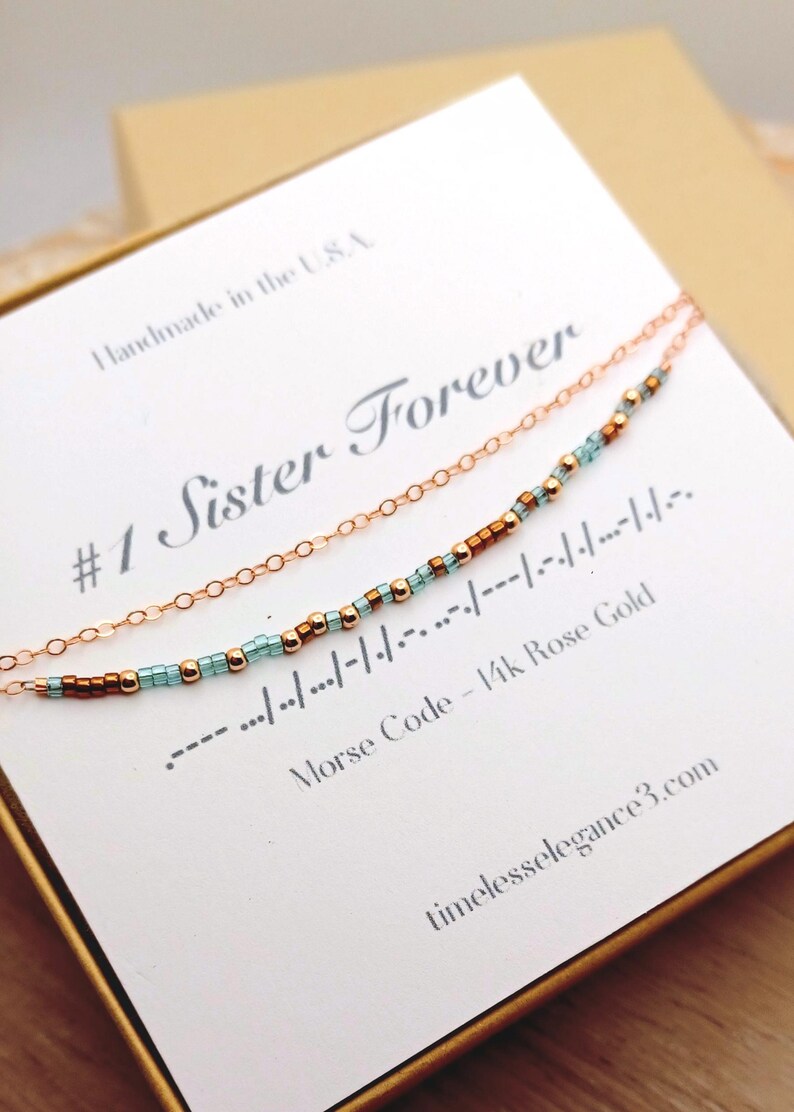 May include: A rose gold necklace with a delicate chain and a beaded section spelling out a message in Morse code. The card reads "#1 Sister Forever" and "Morse Code - 14k Rose Gold". The words "Handmade in the USA" are also visible.