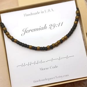 May include: A beaded necklace with black and gold beads, presented in a gift box. The card inside reads "Jeremiah 29:11" and includes Morse code. The text "Handmade in U.S.A." and a website address are also visible.