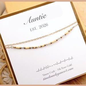 May include: A gold-colored layered necklace with a beaded strand and a delicate chain. The necklace is displayed on a white card with the word "Auntie" and "EST. 2026" printed on it. The card also includes Morse code and the website address timelesselegance8.com.