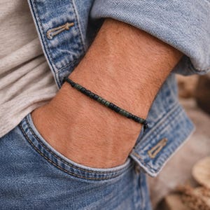 May include: A thin bracelet featuring a repeating pattern of small, cylindrical matte black and dark olive green beads strung on a dark cord. The beads are arranged in a simple, uniform sequence, creating a subtle and understated accessory.