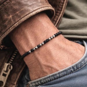 May include: A beaded bracelet featuring a repeating pattern of small, matte black cylindrical beads and polished silver-toned spherical beads. The bracelet is constructed with a thin, flexible cord and maintains a consistent, uniform width throughout.