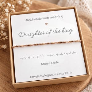 May include: A rose gold beaded bracelet featuring Morse code patterns, presented on a white card inside a brown box. The card displays the text "Handmade with meaning", "Daughter of the king", "Morse code", and "timelesselegance3.etsy.com".