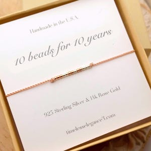 May include: A rose gold bracelet with ten small beads strung on a delicate cord. The bracelet is presented in a beige box with a card that reads "10 beads for 10 years" and "Handmade in the U.S.A."