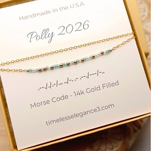 May include: A delicate gold-filled necklace with a Morse code design. The necklace features small beads in gold, clear, and light blue. The card reads "Polly 2026" and "Morse Code - 14k Gold Filled." The item is handmade in the U.S.A.