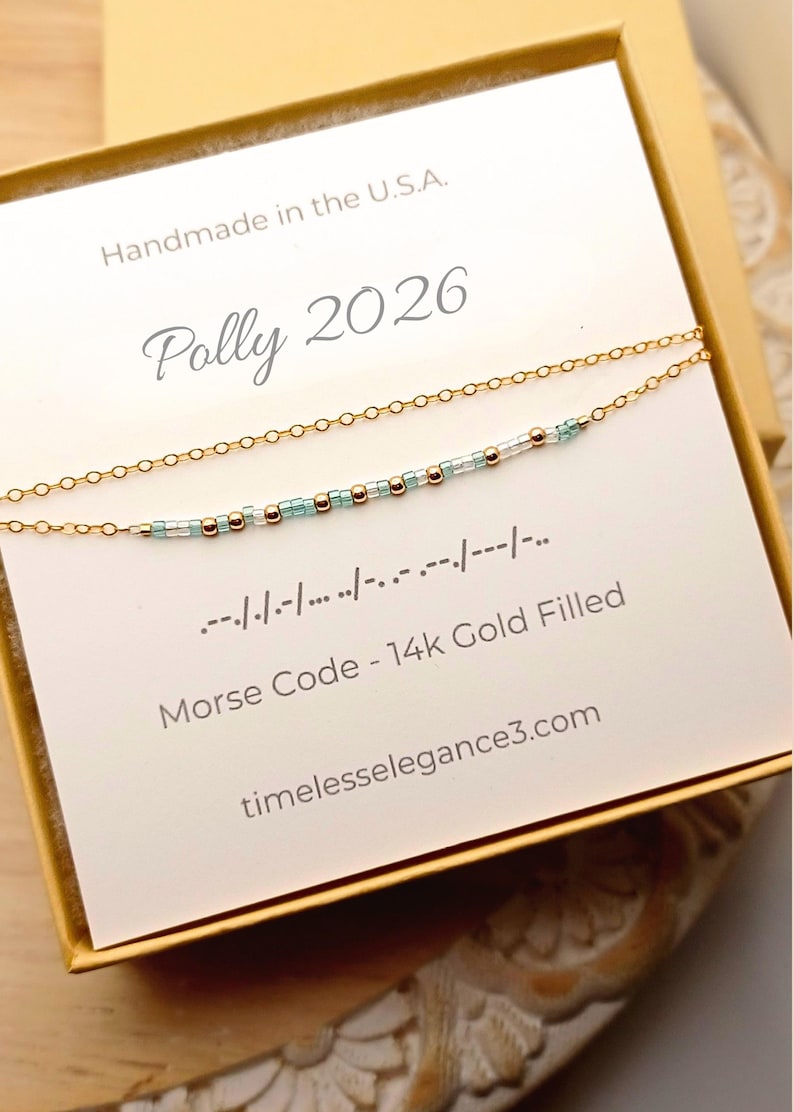 May include: A delicate gold-filled necklace with a Morse code design. The necklace features small beads in gold, clear, and light blue. The card reads "Polly 2026" and "Morse Code - 14k Gold Filled." The item is handmade in the U.S.A.
