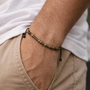 May include: A beaded bracelet with brown and bronze-colored beads. The bracelet is on a brown cord and is worn on the wrist. The person is wearing a white t-shirt and khaki pants.
