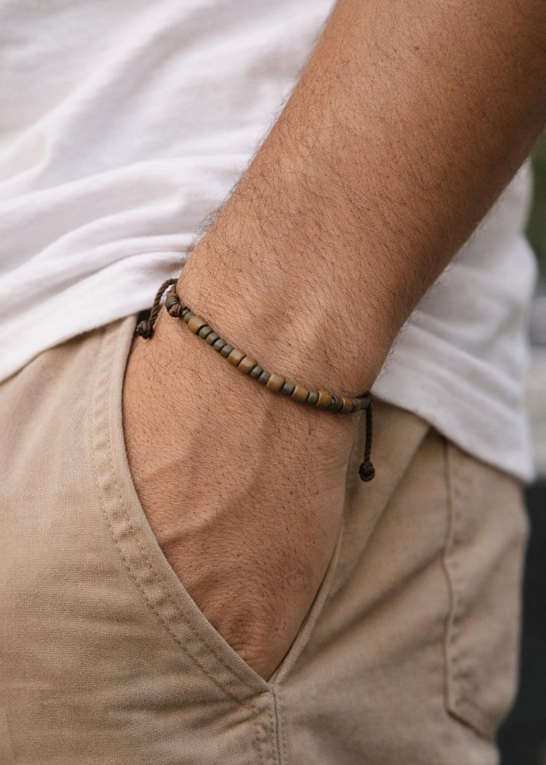 May include: A beaded bracelet with brown and bronze-colored beads. The bracelet is strung on a dark brown cord and is worn on a person's wrist. The person is wearing a white t-shirt and khaki pants.