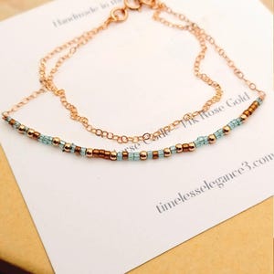 May include: A delicate rose gold necklace with a beaded strand of turquoise and copper-colored beads. The necklace is layered with a fine chain. The card behind the necklace reads "Handmade in the..." and "14K Rose Gold".