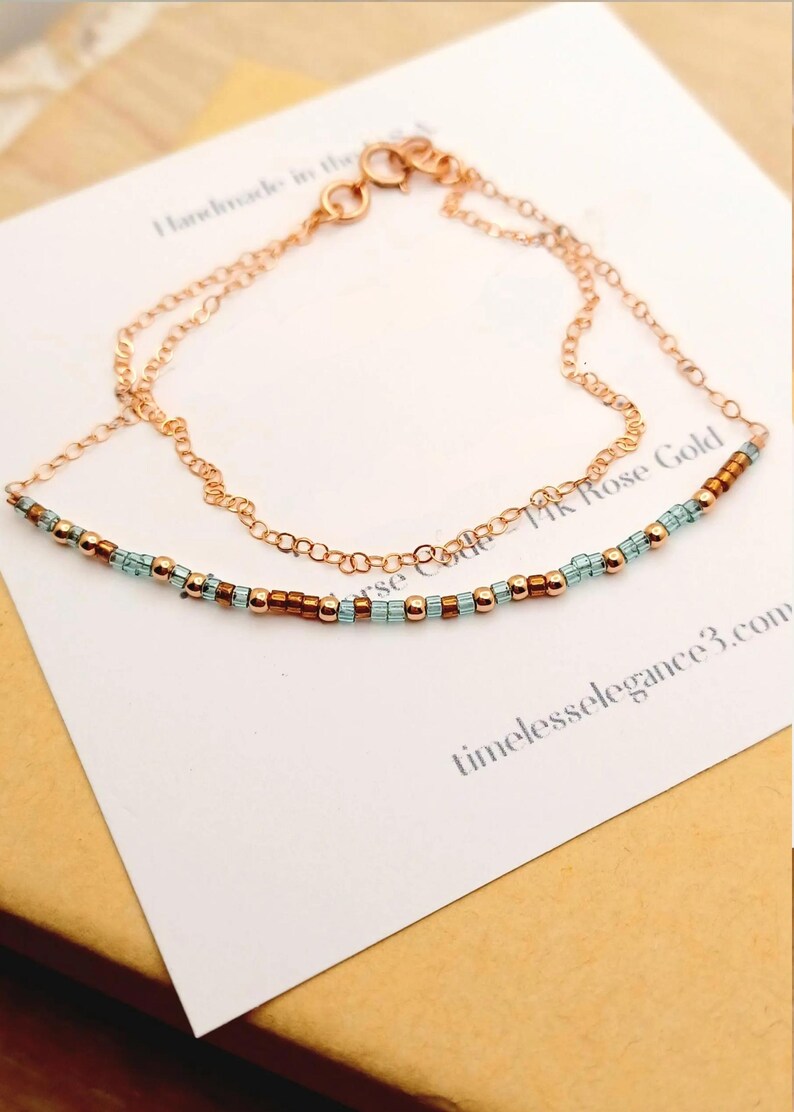 May include: A delicate rose gold necklace with a beaded strand of turquoise and copper-colored beads. The necklace is layered with a fine chain. The card behind the necklace reads "Handmade in the..." and "14K Rose Gold".