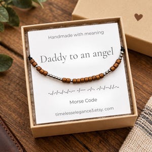 May include: A Morse code bracelet featuring alternating brown wooden beads and small silver metallic beads on a black cord, presented on a white card with the text Daddy to an angel, Morse Code, and the URL timelesselegance3.etsy.com inside a box.