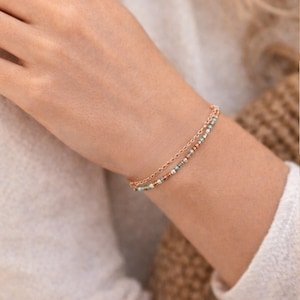 May include: A delicate, double-strand bracelet in rose gold. One strand is a fine chain, the other is adorned with small, colorful beads in shades of turquoise, white, and copper. The bracelet is worn on a wrist.