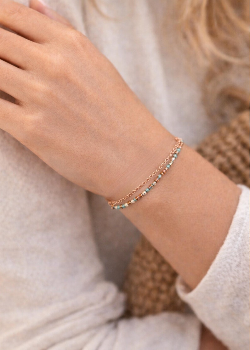May include: A delicate, double-strand bracelet in rose gold. One strand is a fine chain, the other is adorned with small, colorful beads in shades of turquoise, white, and copper. The bracelet is worn on a wrist.