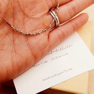 May include: Rose gold necklace with a beaded design, held in a hand. The necklace has two delicate chains and a row of small beads. A card with Morse code and the text "Morse Code - 14k Rose Gold" is visible.