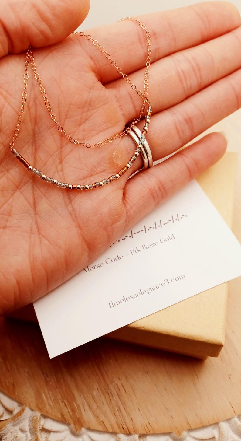 May include: Rose gold necklace with a beaded design, held in a hand. The necklace has two delicate chains and a row of small beads. A card with Morse code and the text "Morse Code - 14k Rose Gold" is visible.