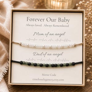 May include: Two Morse code bracelets in a gift box. The top bracelet has white beads on a light cord with the text Mom of an angel. The bottom bracelet has dark beads on a black cord with the text Dad of an angel. The card reads Forever Our Baby.